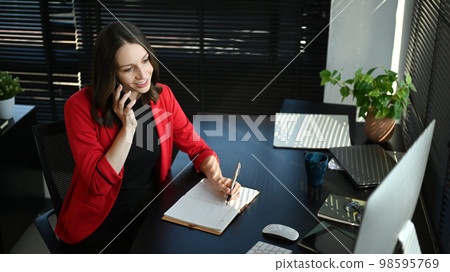 Attractive caucasian businesswoman in elegant red suit having pleasant conversation while sitting at workstation Attractive caucasian businesswoman in elegant red suit having pleasant conversation while sitting at workstation 98595769