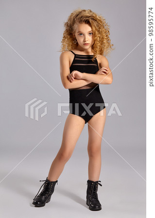 Beautiful little girl gymnast in a black sports swimsuit and boots on a gray background. Beautiful little girl gymnast in a black sports swimsuit and boots on a gray background. 98595774