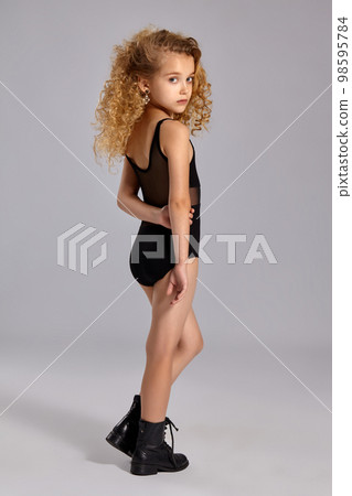 Beautiful little girl gymnast in a black sports swimsuit and boots on a gray background. 98595784