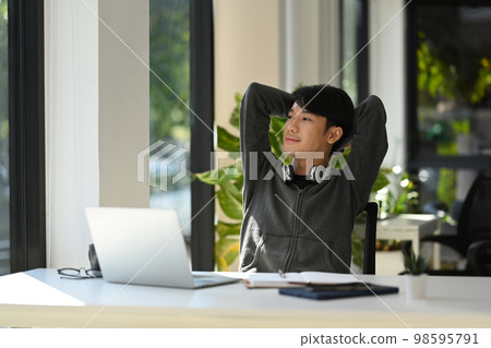 Relaxed young asian man worker sitting back in office chair with hands clasped behind head and looking through window 98595791
