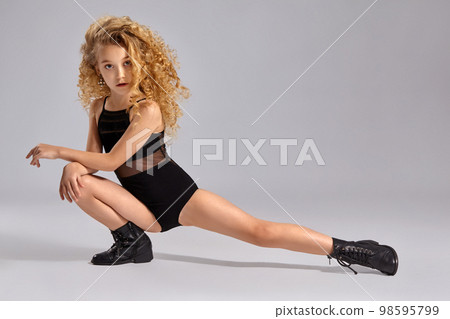 Beautiful little girl gymnast in a black sports swimsuit and boots on a gray background. Beautiful little girl gymnast in a black sports swimsuit and boots on a gray background. 98595799