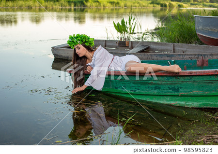 The nymph with long dark hair in a white vintage dress sitting in a boat in the middle of the river. 98595823