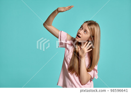 Studio portrait of a beautiful girl blonde teenager in a pink t-shirt posing over a blue background. 98596641