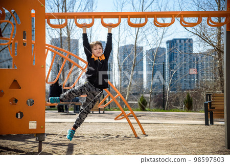 Cute boy is climbing on the playground in the schoolyard. He has a very happy face and enjoy this adventure sports alone outdoor. Warm sunny day 98597803