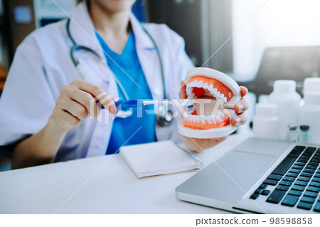 Dentist sitting at table with jaw samples tooth model and working with tablet and laptop in dental office professional dental clinic. 98598858