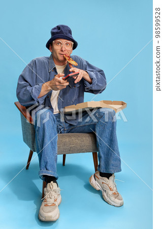 Into game. Cheerful man in jeans clothes sitting with pizza and playing retro game console over blue studio background. Concept of emotions, lifestyle, hobby 98599528