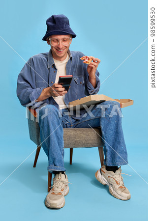 Delightful. Cheerful man in jeans clothes sitting with pizza and playing retro game console over blue studio background. Concept of emotions, lifestyle, hobby 98599530
