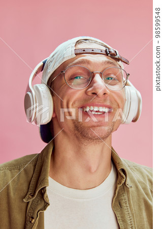 Delightful. Young positive man in glasses, smiling, listening to music in headphones, posing over pink studio background. Concept of emotions, lifestyle Delightful. Young positive man in glasses, smiling, listening to music in headphones, posing over pink studio background. Concept of emotions, lifestyle 98599548