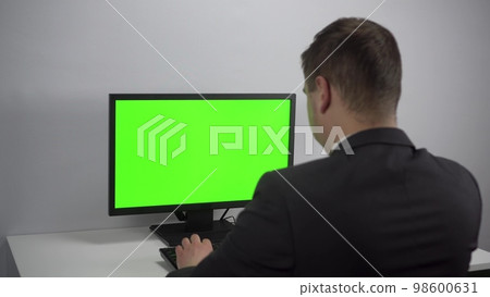 A young businessman is sitting in front of a computer with a green screen. A man in a suit works for a PC. Screen with chroma key. 98600631