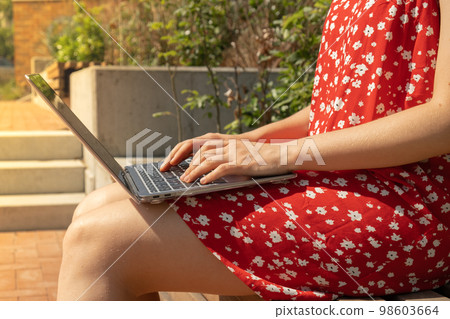 Unrecognizable Young woman in red dress working on laptop. Freelancer's place of work. Study and work online. Remote business education. View webinar. E-learning Workstation on wooden bench. 98603664