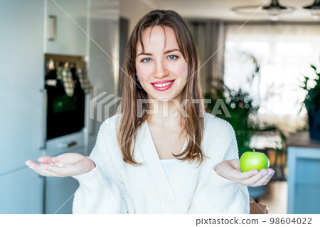 Portrait of young smiling woman holding green fresh apple in one hand, pill, vitamins in another, trying to decide which choice is the best one stand on kitchen. Diet modification, drugs alternative. Portrait of young smiling woman holding green fresh apple in one hand, pill, vitamins in another, trying to decide which choice is the best one stand on kitchen. Diet modification, drugs alternative. 98604022