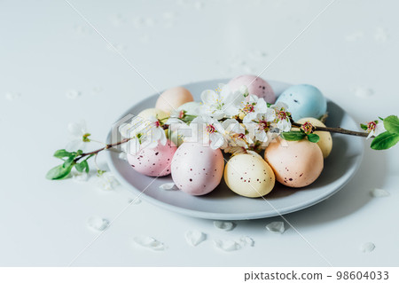 Colored Easter eggs and blooming tree branch on the gray plate with flower petals on the white table. Happy Easter and springtime card. Minimalism and simplicity. Selective focus. Copy space. Colored Easter eggs and blooming tree branch on the gray plate with flower petals on the white table. Happy Easter and springtime card. Minimalism and simplicity. Selective focus. Copy space. 98604033