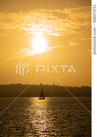 Calm sea with sunset sky and sun through the clouds over. Sailboat in the sea in the evening sunlight over beautiful big mountains background Calm sea with sunset sky and sun through the clouds over. Sailboat in the sea in the evening sunlight over beautiful big mountains background 98605931