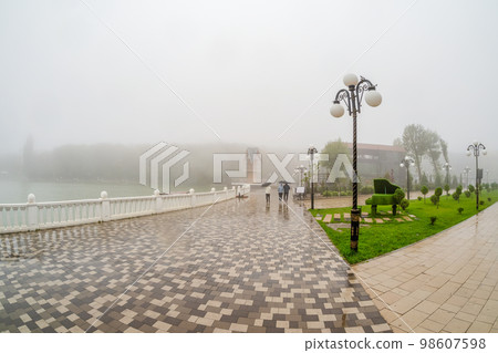 Beautiful wet and foggy embankment with people walking under umbrellas. Foggy rainy autumn landscape with modern park in Zheleznovodsk. Beautiful wet and foggy embankment with people walking under umbrellas. Foggy rainy autumn landscape with modern park in Zheleznovodsk. 98607598