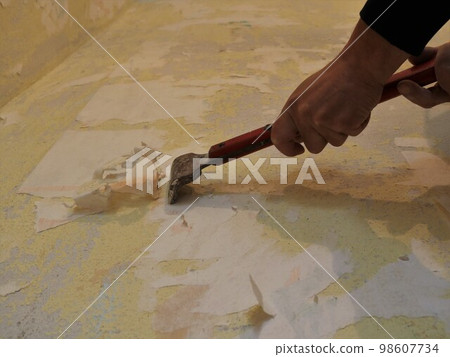 male hands are scraping a surface with the remnants of old peeled wallpaper with a narrow sharp tool on a long handle, casting a shadow on the wall, working with a scraper to clean the surface male hands are scraping a surface with the remnants of old peeled wallpaper with a narrow sharp tool on a long handle, casting a shadow on the wall, working with a scraper to clean the surface 98607734