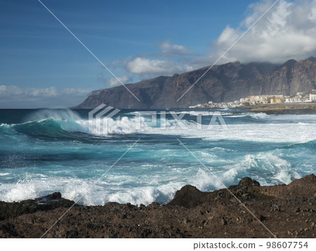 Big breaking waves splash against black lava rock coast. Turquoise blue Atlantic ocean with view of Los Gigantes cliffs and village. Tenerife, Canary islands, Spain. Sunny winter day. Big breaking waves splash against black lava rock coast. Turquoise blue Atlantic ocean with view of Los Gigantes cliffs and village. Tenerife, Canary islands, Spain. Sunny winter day. 98607754