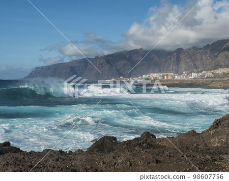 Big breaking waves splash against black lava rock coast. Turquoise blue Atlantic ocean with view of Los Gigantes cliffs and village. Tenerife, Canary islands, Spain. Sunny winter day. 98607756