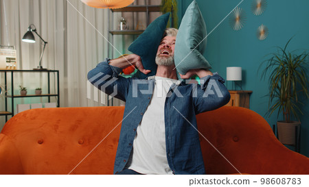 Repair work at neighbours concept, irritated man at home cover ears with pillows annoyed by noise Repair work at neighbours concept, irritated man at home cover ears with pillows annoyed by noise 98608783
