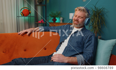 Happy mature man in wireless headphones relaxing sitting on sofa at home listening favorite music 98608789