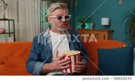 Man sitting on couch eating popcorn and watching interesting TV serial, sport game online at home 98608814