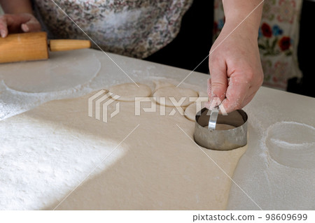 Raw homemade dough for donuts is being cut into round pieces with help of rolling pin on baking table Raw homemade dough for donuts is being cut into round pieces with help of rolling pin on baking table 98609699