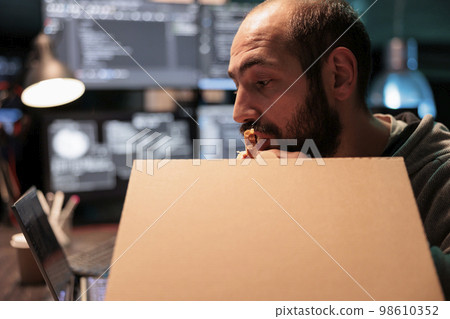 Database coder eating pizza from takeaway delivery, taking bite of food while he works after hours on source code development. Programmer having late dinner with takeout meal in carton box. Database coder eating pizza from takeaway delivery, taking bite of food while he works after hours on source code development. Programmer having late dinner with takeout meal in carton box. 98610352