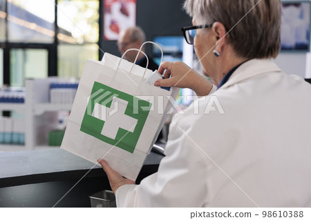 Senior drugstore worker standing at pharmacy counter putting prescription treatment in paper bag preparing package for client. Customers looking at shelves full with medication, buying pills 98610388
