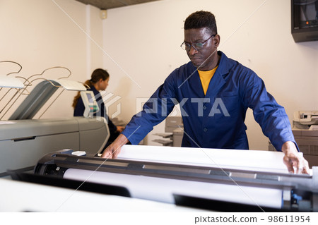 Focused African American male in a blue robe uniform using plotter during workflow in the typography Focused African American male in a blue robe uniform using plotter during workflow in the typography 98611954