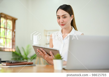Charming millennial Asian businesswoman sits at her desk with a digital tablet Charming millennial Asian businesswoman sits at her desk with a digital tablet 98615703