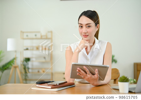 Determined Asian woman sits at the table with her digital tablet, hand on chin Determined Asian woman sits at the table with her digital tablet, hand on chin 98615705