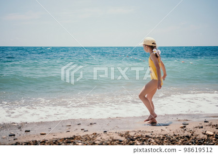 Cute little girl running along the seashore against a clear blue sea and rejoices in the rays of the summer sun. Beautiful girl in yellow swimsuit running and having fun on tropical beach. 98619512
