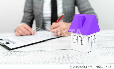 Businesswoman sitting at table and writing in notebook. olored paper house standing on desk. Display important information. Executive Display the latest news. 98620644