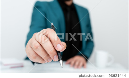Male model in Blue suit sitting at white table And Pointing With Pen On Important Message. Gentleman Showing Critical Announcement. Coffee cup on deck. 98621444