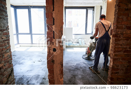 Back view of male worker using troweling machine while screeding floor in apartment under renovation. Man finishing concrete surface with floor screed grinder machine in room with large windows. 98623173