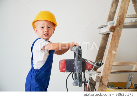 Adorable little boy construction worker holding electric power drill and looking at camera. Cute child wearing work overalls and safety helmet while standing on ladder against white wall. 98623175
