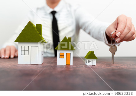 Businesswoman sitting at table holding keys. olored paper houses standing on desk. Display important information. Display the latest news. Critical Data Shown. 98623338