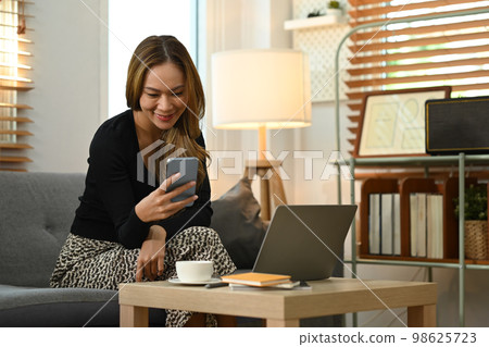Joyful youngwoman using smart phone chatting with friends in social network while sitting on couch at home Joyful youngwoman using smart phone chatting with friends in social network while sitting on couch at home 98625723