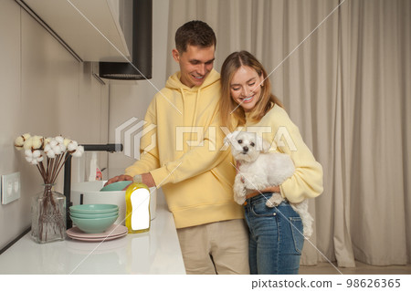 Portrait of happy couple in yellow hoodies, man washing the dishes, woman holding little white dog Portrait of happy couple in yellow hoodies, man washing the dishes, woman holding little white dog 98626365