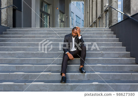 Upset african american man sitting on stairs outside office building, businessman bankrupt in despair, mature boss depressed in business suit outdoors. 98627380