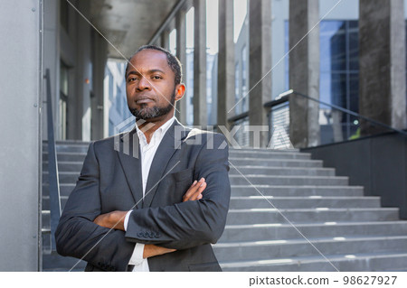 Serious thinking businessman with crossed arms looking at camera, portrait of african american investor boss outdoors in office, man in business suit. 98627927
