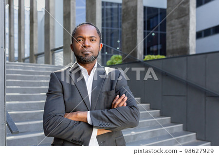 Serious thinking businessman with crossed arms looking at camera, portrait of african american investor boss outdoors in office, man in business suit. Serious thinking businessman with crossed arms looking at camera, portrait of african american investor boss outdoors in office, man in business suit. 98627929