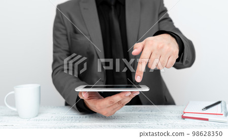 Businessman in gray jacket sitting at table holding mobile phone And Pointing With One Finger On Important Message. Gentleman Showing Critical Announcement. Businessman in gray jacket sitting at table holding mobile phone And Pointing With One Finger On Important Message. Gentleman Showing Critical Announcement. 98628350