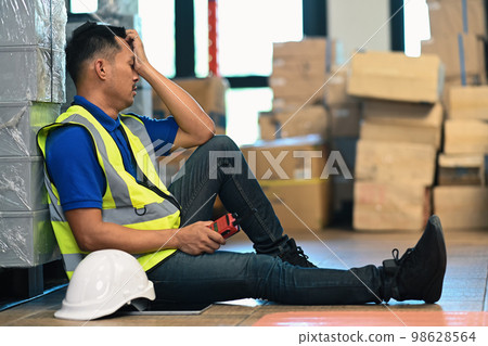 Tired man warehouse worker in jacket leaning against metallic rack while having short break Tired man warehouse worker in jacket leaning against metallic rack while having short break 98628564
