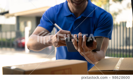 Cropped shot of delivery man in blue uniform using digital tablet while standing at open van full of delivering package Cropped shot of delivery man in blue uniform using digital tablet while standing at open van full of delivering package 98628600