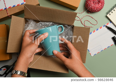 Overhead view of woman putting ceramic cup into cardboard box, preparing to pack gifts for holiday season 98628646