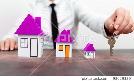 Businesswoman sitting at table holding keys. olored paper houses standing on desk. Display important information. Display the latest news. Critical Data Shown. 98629329