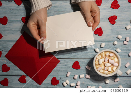 Female hands holding empty valentine card mock up red envelope with white cup of coffee and marshmallows on wooden blue background. Romantic Small hearts Valentine day. Blank paper card copy space for Female hands holding empty valentine card mock up red envelope with white cup of coffee and marshmallows on wooden blue background. Romantic Small hearts Valentine day. Blank paper card copy space for 98630519