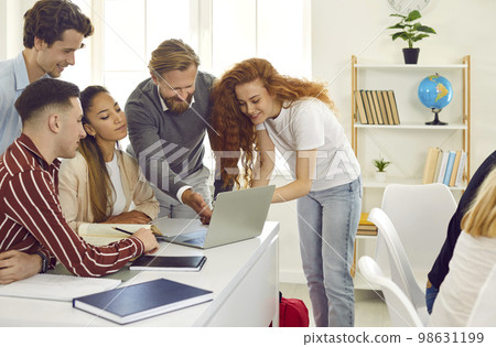 Professor and students are working on project on laptop together sitting at desk and standing near. Professor and students are working on project on laptop together sitting at desk and standing near. 98631199