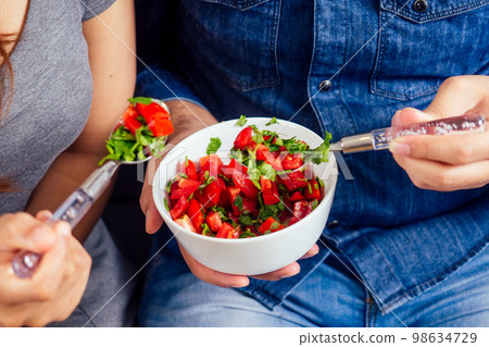 Young indian couple sitting in they living room and eating vegetable salad Young indian couple sitting in they living room and eating vegetable salad 98634729
