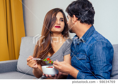 Young indian couple sitting in they living room and eating vegetable salad Young indian couple sitting in they living room and eating vegetable salad 98634769